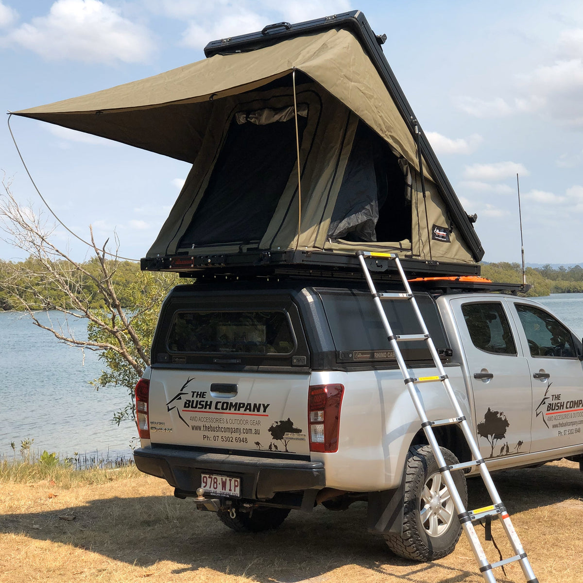 Bush Company AX27 clamshell rooftop tent open on a canopied Isuzu D-Max by a lake, with the ladder down