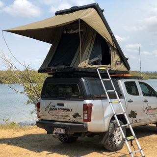 Bush Company AX27 clamshell rooftop tent open on a canopied Isuzu D-Max by a lake, with the ladder down