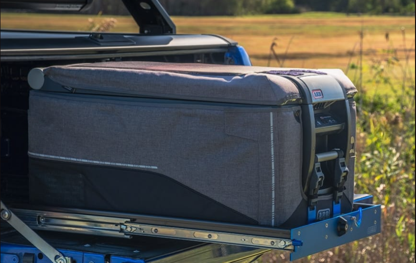An ARB fridge in its padded grey cover strapped to a slide, pulled out over the tailgate of a ute with a paddock behind