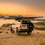 White SUV with a extended roof rack and spare tire on a beach at sunset with Destination 4WD 270 Degree Freestanding Awning