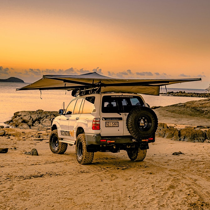 White SUV with a extended roof rack and spare tire on a beach at sunset with Destination 4WD 270 Degree Freestanding Awning