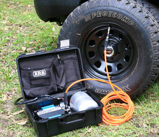 ARB twin piston compressor in its open travel case beside a vehicle on grass, the orange air hose connected to a Pro Comp wheel on a mud terrain tyre
