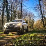 White Mercedes-Benz SUV driving on a forest path with trees and grass on either side.