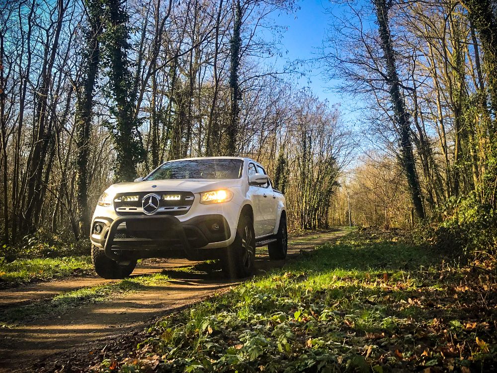 White Mercedes-Benz SUV driving on a forest path with trees and grass on either side.