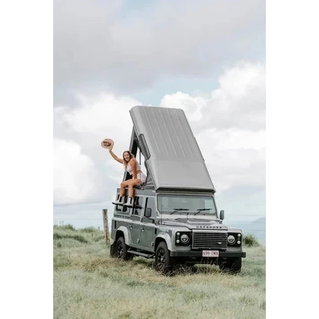 Person sitting on a Land Rover Defender with an open roof in a grassy field.