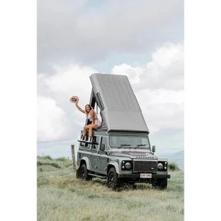 Person sitting on a Land Rover Defender with an open roof in a grassy field.