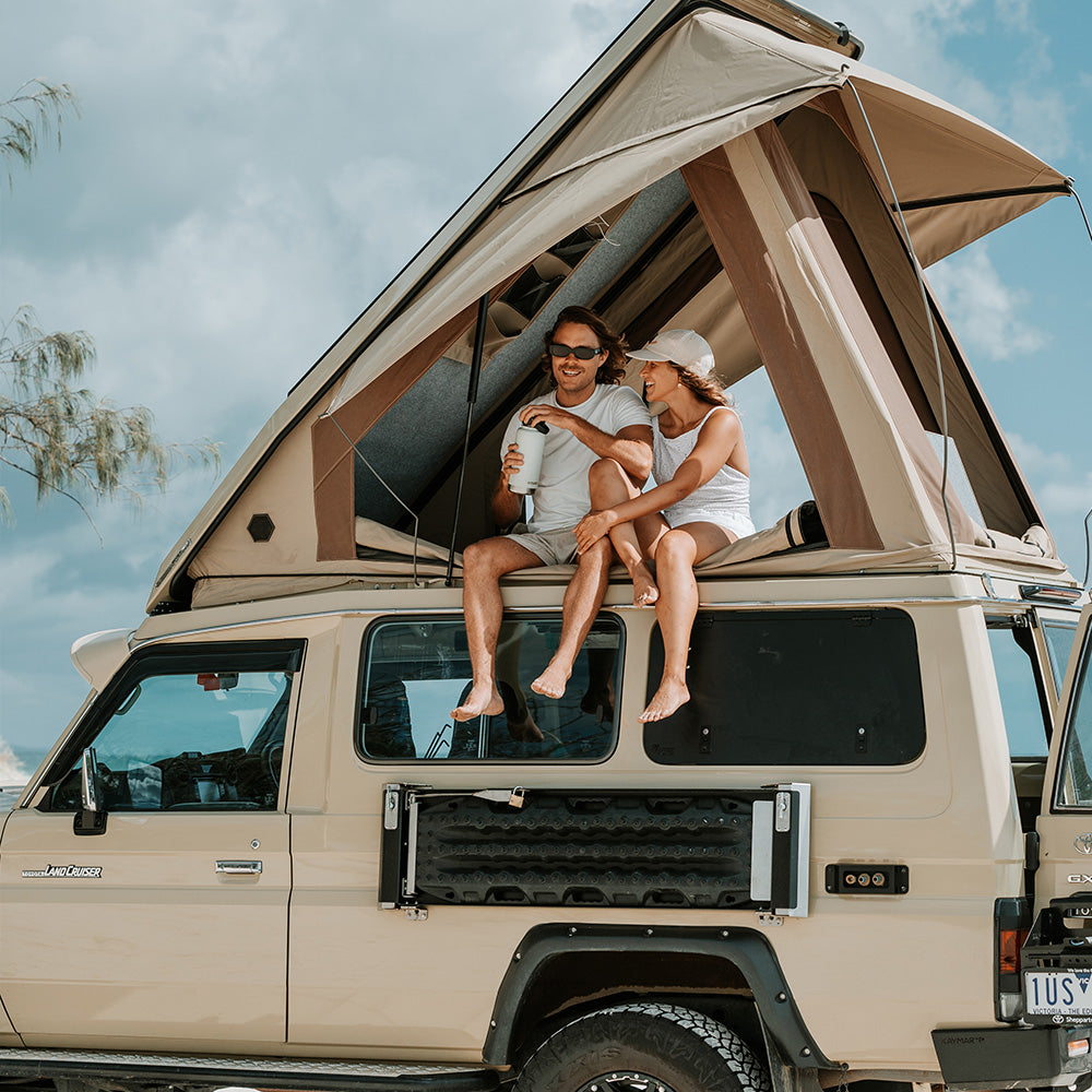 Two people sitting in the raised Headspace roof of a beige Troopy against a blue sky