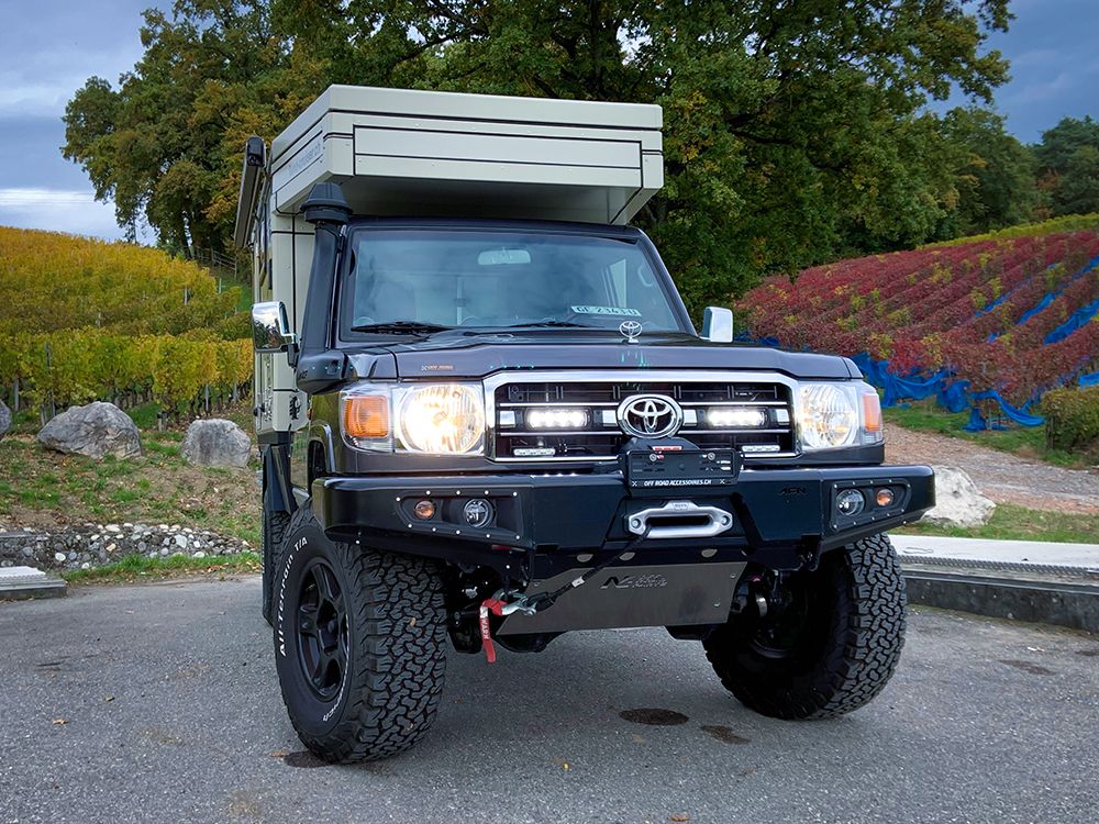 Black Toyota truck with a camper shell on a road with trees and colorful fields in the background