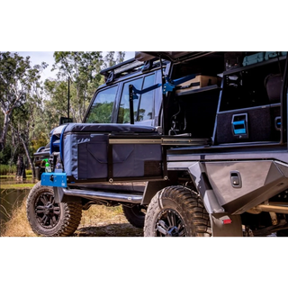 A Toyota LandCruiser troop carrier parked by water with the rear open, a fridge in a padded cover sitting out on the MSA drop slide and a canvas awning arm folded above it