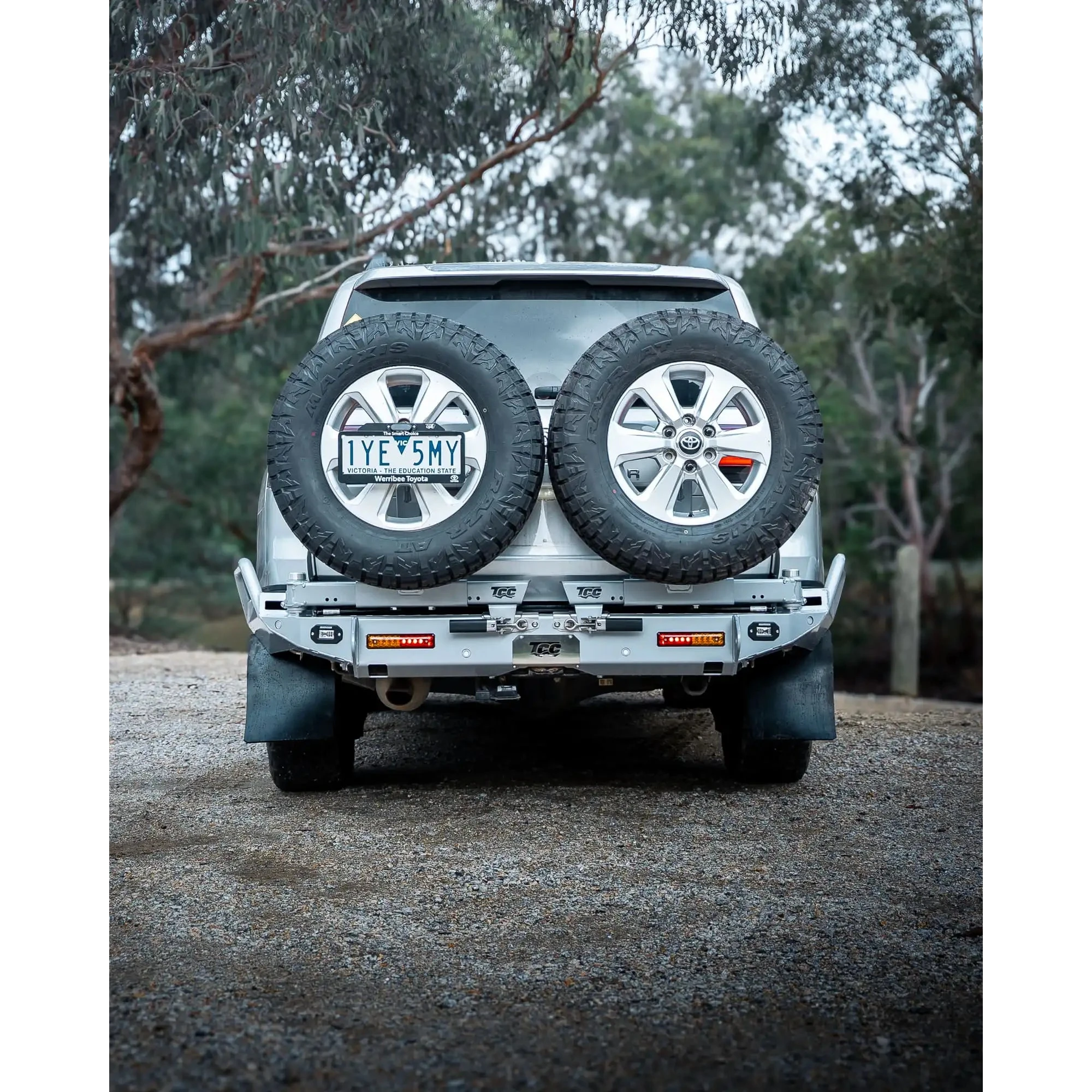 Back view of a silver SUV with spare tires on a gravel road surrounded by trees – TCC Toyota LC300 Series Rear Bar