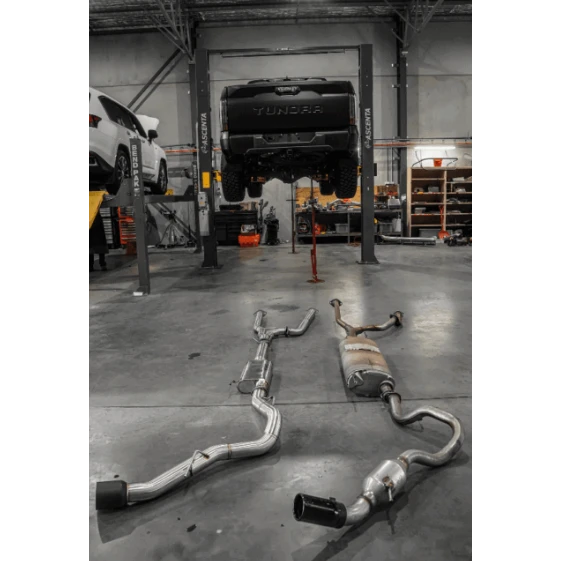 Exhaust system on the floor of a garage with a vehicle on a lift in the background – Torqit 3″ CAT Back Performance EXHAUST Toyota Tundra 3.5L V6 Petrol