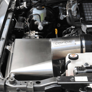 Torqit stainless airbox installed in an engine bay, the brushed panel carrying the Torqit name with the intake hose clamped to the outlet and the brake reservoir and coolant bottle visible alongside