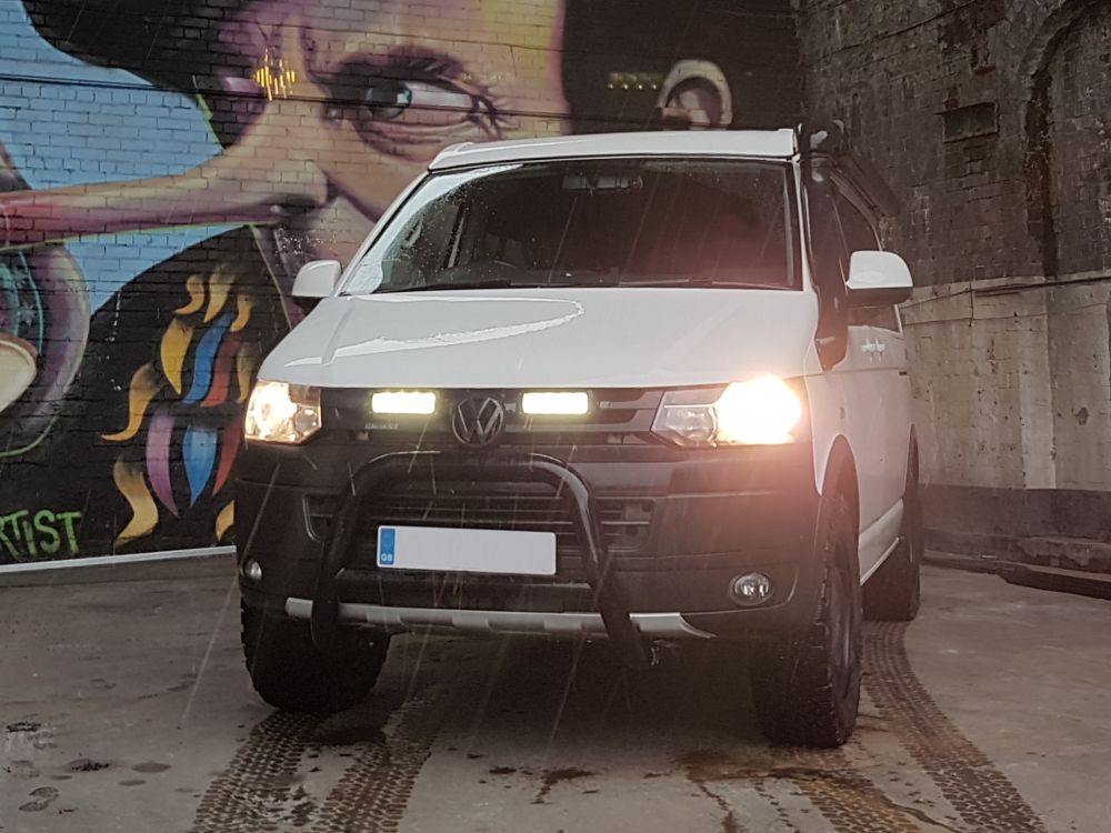 White Volkswagen van with headlights on in front of a colorful mural with Lazer Lamps Grille Kit for Volkswagen T5.1