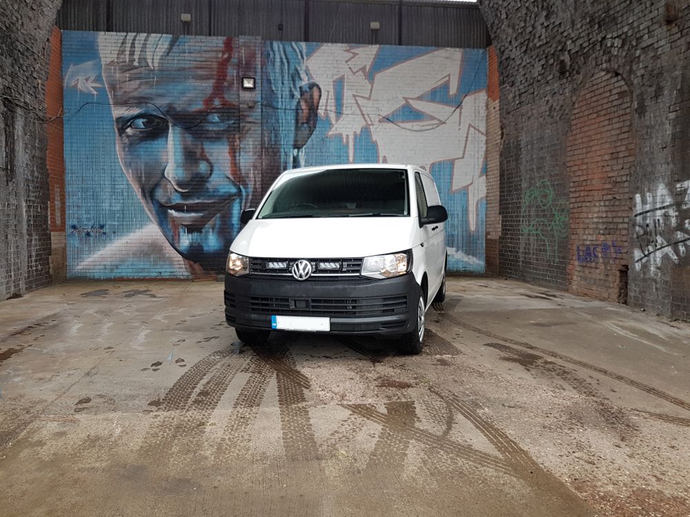 White van parked in front of a mural on a brick wall.